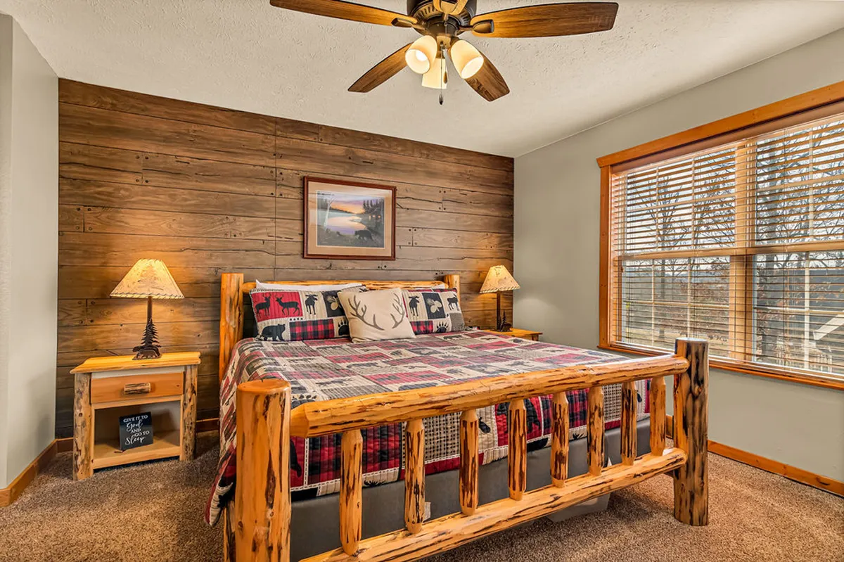 A comfortable loft bedroom with a wooden bed frame, plaid bedding, nightstand with lamp, and framed artwork. The space feels private yet connected to the rest of the cabin, perfect for restful sleep after a day of adventure.