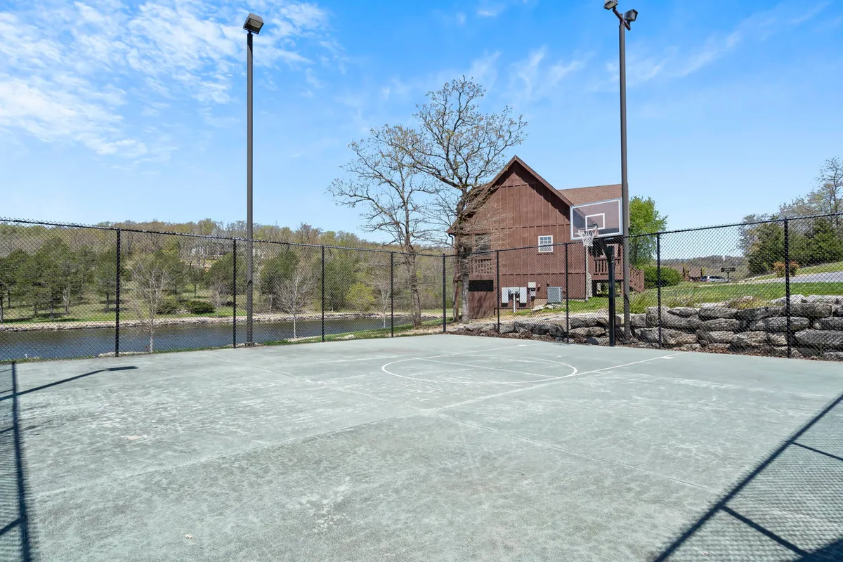 An energetic outdoor basketball court bordered by tall trees and offering a partial lake view, with a rustic cabin-like structure nearby. The space is ideal for family basketball games, picnics, or a stroll along the nature trail.