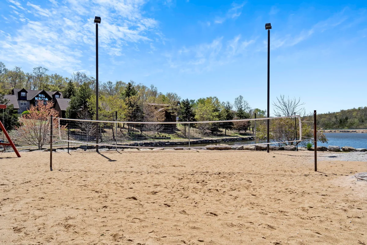 An outdoor sand volleyball court set beside a tranquil body of water, bordered by trees and surrounded by natural beauty. The area reflects a recreational, social atmosphere perfect for both active play and relaxation along the shoreline.