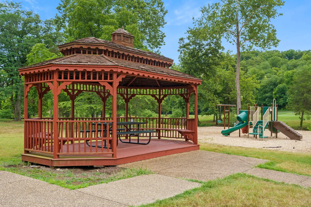 A cheerful playground scene featuring a gazebo, modern playground equipment, and a paved pathway. Children can be seen playing, while families gather nearby for picnics or socializing, all set against a backdrop of trees and open sky.