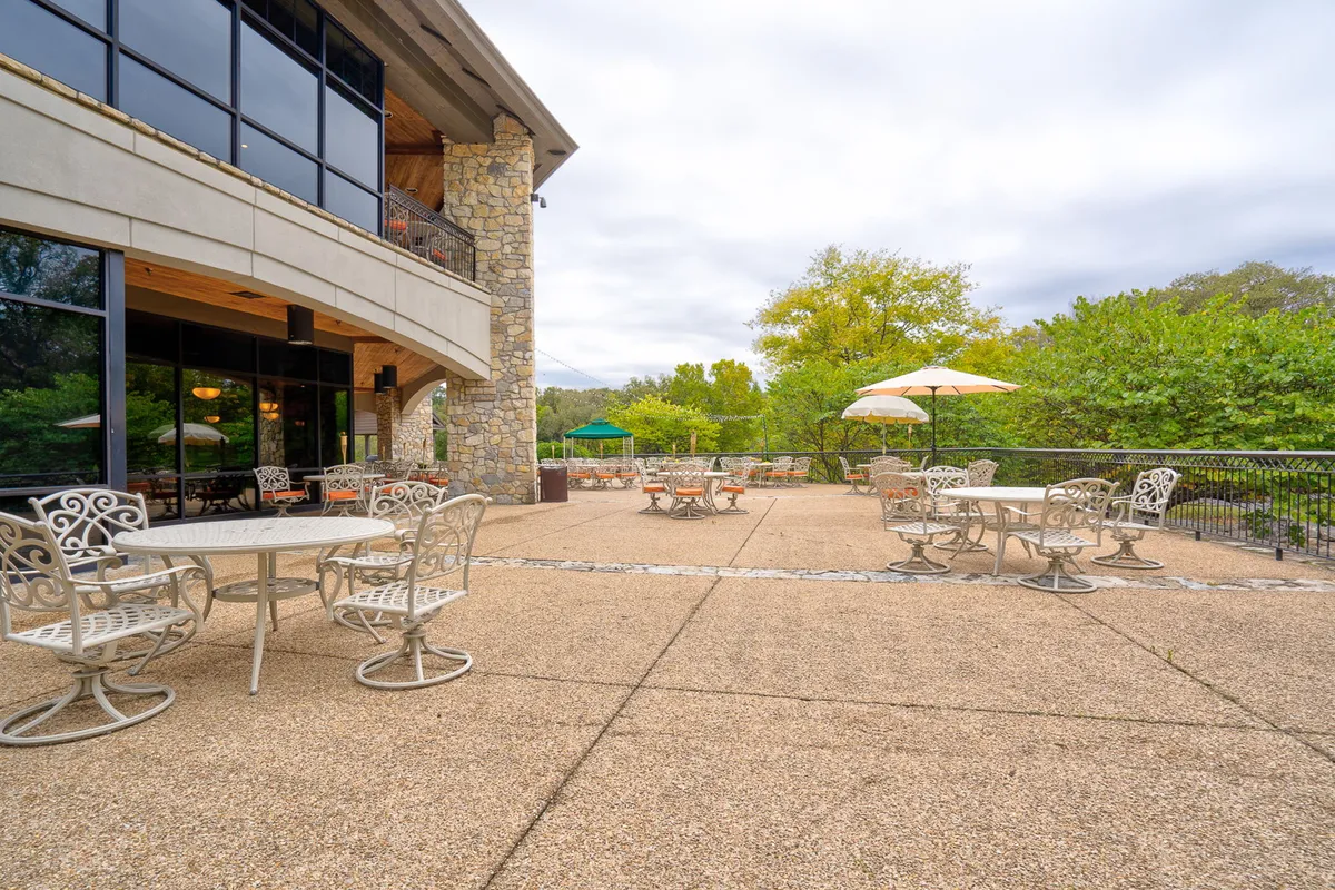 A charming outdoor dining and lounge area with stone walls, cozy seating beneath large umbrellas, and towering green trees in the background. The space is ideal for socializing, enjoying a meal, or relaxing after a round of golf, with a warm, inviting sunset ambiance.