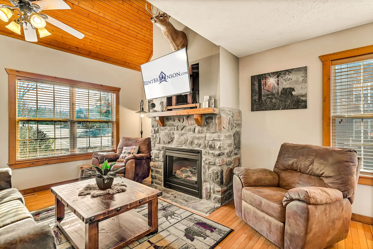 The image showcases a cozy cabin interior featuring a rustic stone fireplace as the centerpiece, a flat-screen television above, vaulted wooden ceilings, and plush seating arranged for relaxation. The atmosphere is warm and inviting, ideal for unwinding or socializing in comfort.