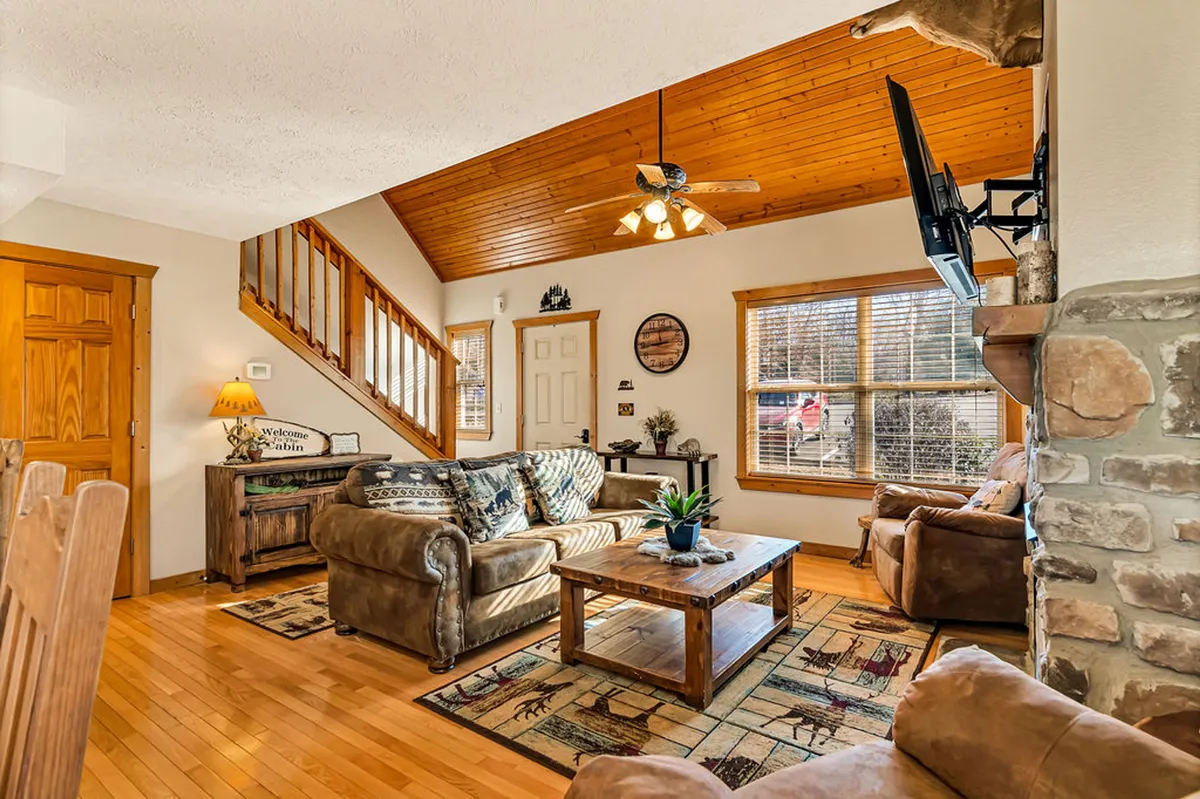 A welcoming living room with a wooden ceiling, stone fireplace, comfortable seating area, coffee table with decorative plants, and inviting wall art. The space is warm and homey, ideal for relaxing and socializing with family or friends.