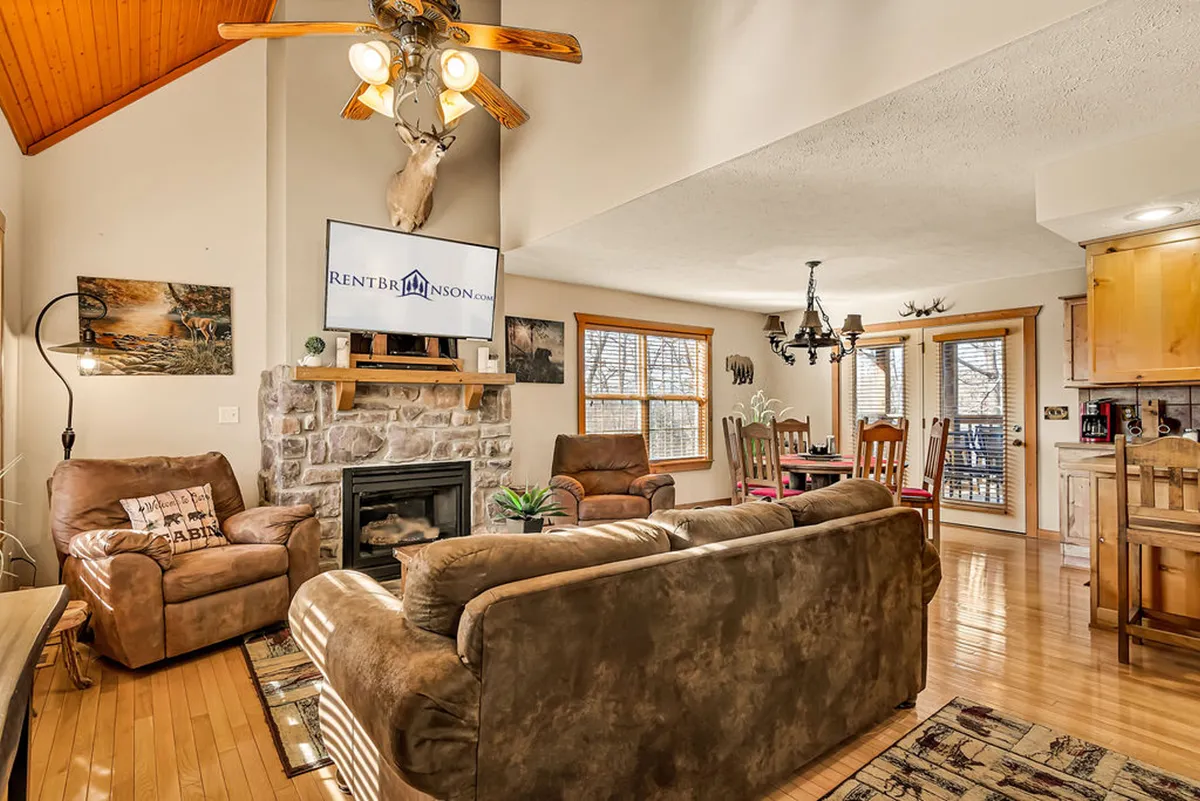 A beautifully styled, open interior showcasing a stone fireplace, wooden ceiling, cozy seating area, dining space, and rustic decorative touches like a mounted animal head. The layout is ideal for both quiet relaxation and group activities such as movie nights or family dinners.