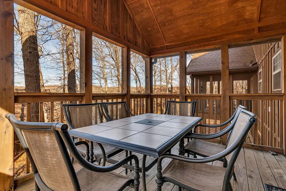 A screened-in outdoor dining patio surrounded by lush trees, with a rustic wooden table and cozy chairs. The space beckons for relaxed desserts or after-dinner conversations, offering a tranquil transition from indoor dining to the beauty of nature and fresh Ozarks air.