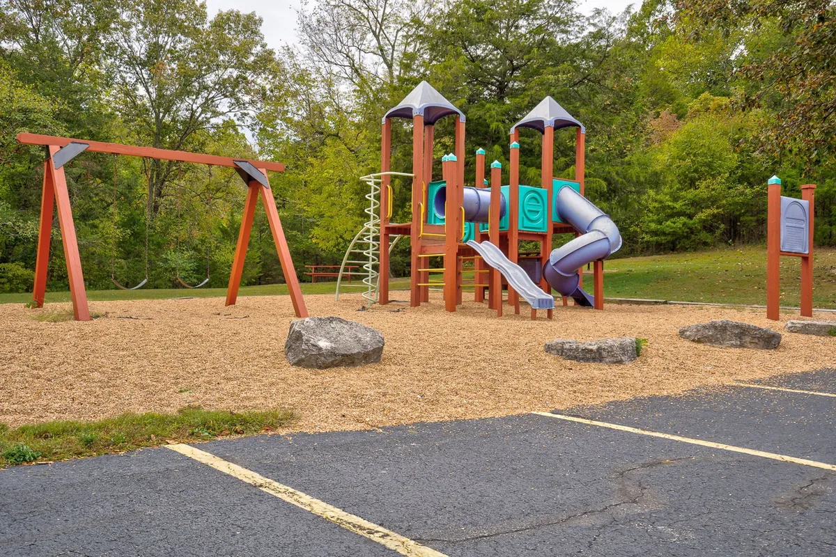 A playful playground scene featuring modern equipment, a swing set, and natural rock features, set amidst trees. Children can be seen enjoying the space, reflecting a family-friendly environment.
