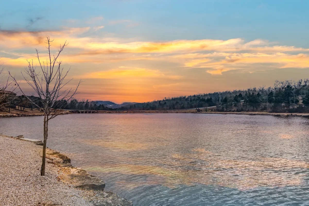 A serene lakeside view at sunset, with soft light illuminating the tranquil water, a solitary tree, and distant rolling hills. The peaceful ambiance evokes the magic of evening strolls or quiet moments by the water.