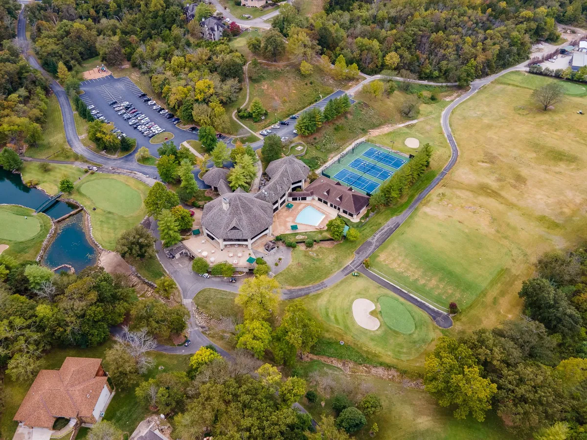 A wide view of the main clubhouse set amid lush greenery, with a sparkling swimming pool, tennis courts, and the golf course visible. The inviting pool is surrounded by loungers, and the clubhouse stands as a social hub for the resort. The landscape conveys the range of outdoor activities and amenities available at StoneBridge Resort.